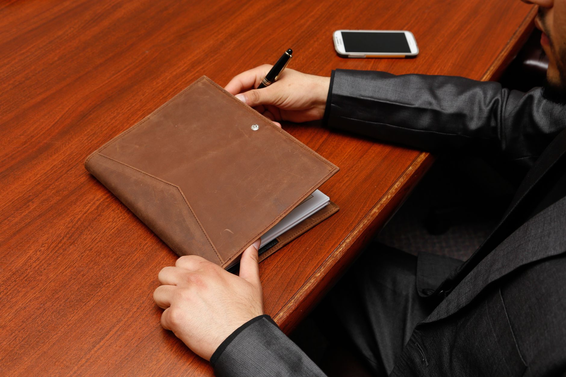 Male executive writing notes during a business meeting using a leather notebook.